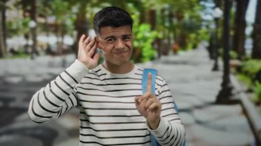 Çizgili gömlekli ve mavi sırt çantalı genç adam şehir sokaklarında bulanık ağaçlar ve arka planda dinleme niyeti taşıyan insanlar.