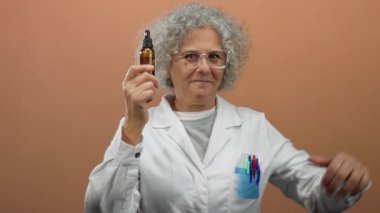 Gri saçlı, gülümseyen ve elinde damlatan şişeyle kendini işaret eden kahverengi duvarlı, beyaz önlüklü ve gözlüklü bir kadın..