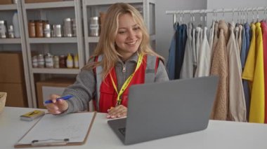 Genç gönüllü bir kadın hayır kurumunda dizüstü bilgisayar kullanıyor, kalemle notlar alıyor, giysi ve bağış eşyalarıyla çevrili, kendini adamışlık ve bağlılık sergiliyor..