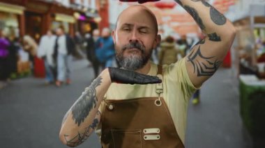 Dövmeli ve sakallı kel adam hareketli şehir sokaklarında kahverengi tulumlu ve siyah eldivenli poz veriyor..