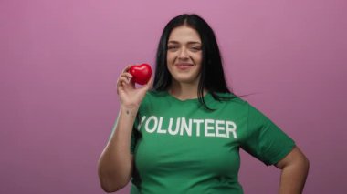 Kırmızı kalp şeklinde bir nesne tutan kadın pembe arka plan aydınlatma ve nezaket karşısında gönüllü gömleği giyerek güvenle gülümsüyor..