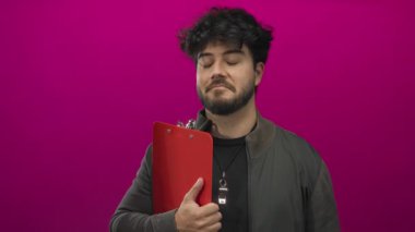 Sakallı genç İspanyol bir adam pembe arka planda pano tutuyor. Ceket ve ıslık çalıyor. Kararlı ve düşünceli görünüyor.