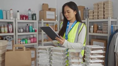Yansıtıcı yelek giyen ve yardım merkezinde tablet tutan bir kadın, bağış kutuları ve yiyecek malzemeleriyle çevrili, gönüllü bir çevrede örgütlenmeyi öneriyor..