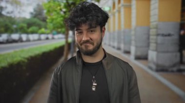 Arka planda ağaçlar ve arabalarla şehir sokağında el çırpan sakallı genç İspanyol bir adam sıradan moda ve doğal ifade sergiliyor..