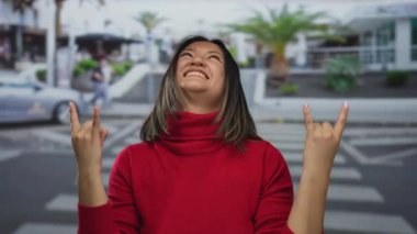 Kırmızı kazaklı genç Çinli kadın taş el hareketi yapıyor, hareketli bir şehir caddesinde neşeyle gülümsüyor, şehir enerjisini ve ruhunu yakalıyor..