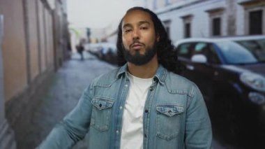 Sakallı ve uzun saçlı bir adam sıkılmış yumruğunu kaldırıyor ve şehir caddesinde park halindeki arabanın yanında kot ceket giyiyor..