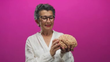 Gri saçlı yaşlı kadın, parlak pembe arka planda beyin modelini inceliyor. Nöroloji ve eğitim bilgisini ve merakını gösteriyor.