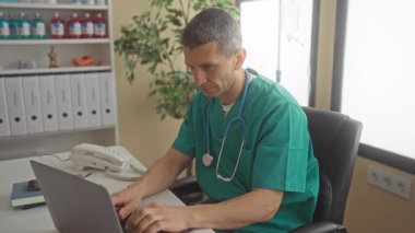 Klinikteki doktor steteskoplu üniforma giyiyor. Etrafı tıbbi dosyalar ve ekipmanlarla çevrili bir dizüstü bilgisayarla çalışıyor. Profesyonel bir sağlık hizmeti ortamı yaratıyor..