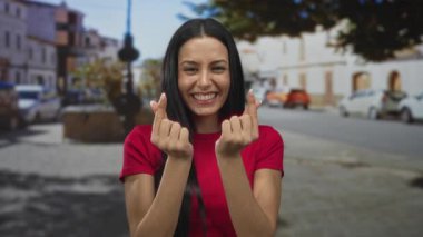 Şehirdeki genç İspanyol kadın parmaklarıyla kalp şekli yapıyor, güneşli bir sokakta arka planda arabalar ve binalarla neşe içinde gülümsüyor..
