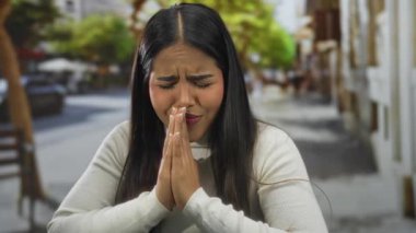 Sokakta gözleri kapalı ve elleri kapalı dua eden kadın, şehir ortamında duyguları ifade ediyor, bulanık ağaçlar ve binalarla çevrili, beyaz süveter giyiyor..