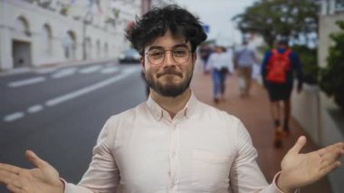 Young man with beard wearing glasses smiles while standing outdoors with hands in a prayer gesture on a busy street