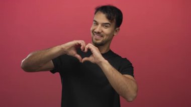 Genç İspanyol adam gülümsüyor ve elleri ile kalbi şekillendiriyor izole edilmiş kırmızı arka plana karşı sevgi ve pozitiflik sergiliyor