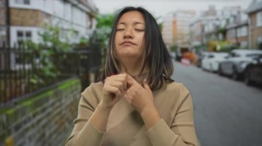 Şehir caddesinde öksüren genç bir kadın, bej bir kazak giyiyor, şehir mimarisi ve park edilmiş arabalarla çevrili, şehir ortamında samimi bir anı yakalıyor..