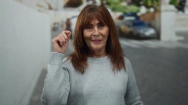 Yaşlı İspanyol bir kadın şehir sokaklarında şakacı bir şekilde bir telefon konuşmasını işaret ediyor ve taklit ediyor. Açık bir şehir ortamında mizah ve bağlantı kuruyor..