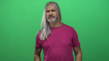 Yeşil stüdyoda uzun gümüş saçlı, elleri kalçasında morumsu gömlekli, yaşlı, dayanıklılık sıkıntısı çeken bir adam..