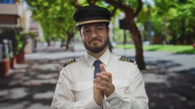 Sakallı genç adam pilot üniforması giyiyor güneşli bir sokakta el çırpıyor..