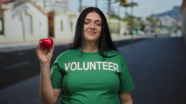 Kıvrımlı genç bir kadın, yeşil bir gömlek giyip, kenarı işaret ederken elinde kırmızı kalp şeklindeki bir nesneyle şehrin caddesinde kendine güvenerek duruyor..