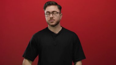 Sakallı genç bir adam, parlak kırmızı bir arkaplanı işaret ediyor. Gözlük takıyor ve siyah bir gömlek giyiyor. Odak ve yönünü izole bir ortamda belirtiyor..