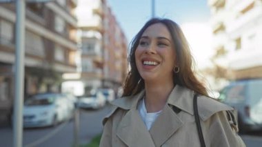 Güneşli bir günde şehir caddesinde gülümseyen bir kadın bej bir ceket giyiyor. Arka planda binalar ve arabalar var. Açık hava yaşam tarzını gözler önüne seriyor..