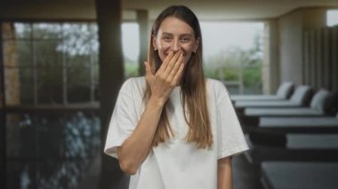 Kadın eli ağızında ve spa binasında kapalı havuz kenarında gülümsüyor, beyaz tişört ve inci küpeler takıyor; neşe dolu neşe dolu..