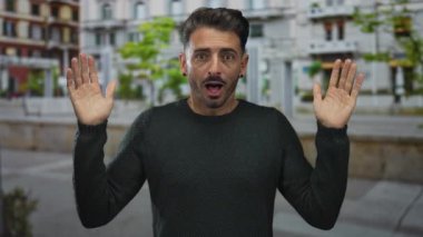 Young hispanic man lifts both hands in a surprised gesture along a busy city street sidewalk; surprise.