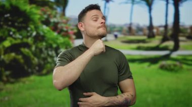 Genç İspanyol bir adam güneşli bir parkta jest yapıyor. Canlı yeşil elbiseler giyiyor ve kolunda dövmeler var. Modern açık hava tarzını ve ifadesini yansıtıyor..