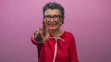 Gri saçlı, ağzı kapalı, pembe arka planda kahkahalar atan, gözlüklü ve kırmızı gömlekli son sınıf öğrencisi bir kadın..