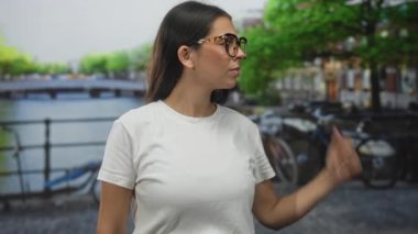 Beyaz tişörtlü genç esmer kadın, bisiklet korkuluklara ve köprü manzarasına kilitlenmiş Amsterdam Kanalı caddesinde kameraya yumruk atıyor..