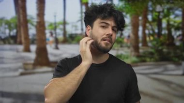 Sokakta sakallı genç bir adam ağaçlarla çevrili yüzüyle güneşli şehir ortamında düşünceli bir ifade veriyor..