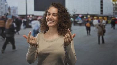 Kalabalık ve reklam panosu arkasında, bej süveter, keyif güveni olan kalabalık şehir sokağında gülümseyen ve başparmağını gösteren bir kadın..