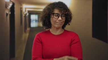 Kirmizi kazak giyen kadin, loş bir otel koridorunda parmaklari duvara dolasiyor; merak icerisi arastirmalar.