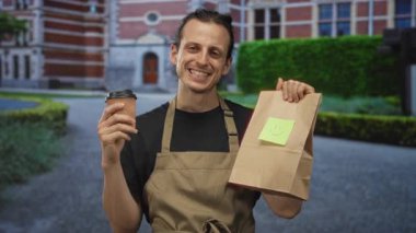 Önlüklü bir adam sağ elinde bir paket kahve fincanı ve tuğla binanın önünde gülümserken yapışkan bir gülümseme notuyla bir kese kağıdı tutuyor..