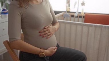 Elleri karnında hamile bir kadın bebek odasında gülümsüyor. Bebek için sevinç ve beklenti içinde.