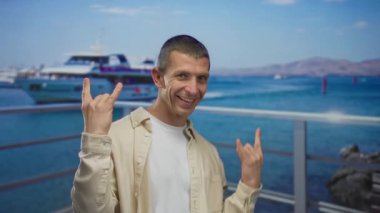 Deniz kıyısında yatın önünde gülümseyen bir adam mavi gökyüzünün altında okyanus ve dağlarla kaya el hareketi yapıyor.