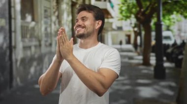 Sokak lambaları ve park etmiş scooterların yakınındaki yapraklı bir caddede dua jestine sarılmış genç İspanyol bir adam..