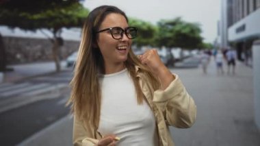 Kadın kenetlenmiş yumruklarını sallıyor ve uzaktan binaları ve yayaları olan bir şehir caddesinde gülümsüyor; zafer başarısı.