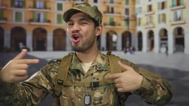 Kamuflaj üniformalı adam, bina cephesi yakınlarında el yazısı ve sırt çantası takarken parmak göğse işaret ediyor..