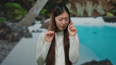 Uzun saçlı ve beyaz gömlekli bir kadın, lüks bir tatil köyünde, bir havuzun yanında, güneşli açık hava ortamındaki yüz ifadelerini vurguluyor..