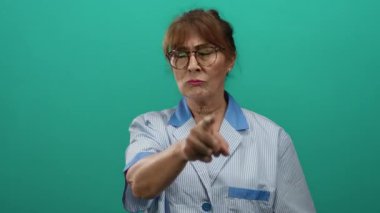 Temizlik üniforması giyen İspanyol kıdemli kadın, yeşil duvar arka planına el hareketleriyle karşı koyuyor..