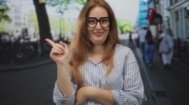 Gözlüklü bir kadın, kollarını önünde çaprazlarken şehir caddesinde ileriyi işaret ediyor..