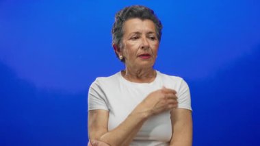 Gri saçlı, beyaz tişörtlü, mavi arka planlı düşünceli yaşlı kadın derin düşüncelere dalmış görünüyor..