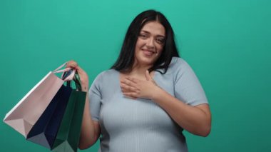 Alışveriş torbaları tutan genç bir kadın yeşil duvara karşı güvenle gülümsüyor, büyük boy moda ve canlı bir geçmişe sahip pozitif alışveriş deneyimi sergiliyor..