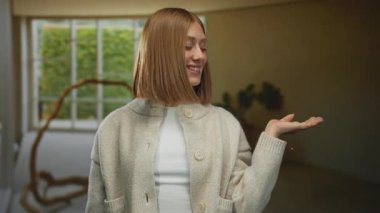 Genç sarışın kadın rahat bir otel odasında gülümsüyor, süveter giyiyor, arka planda manzaralı bahçe manzaralı büyük bir pencerenin yanında duruyor..