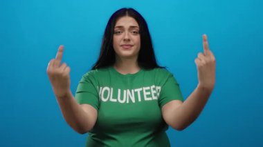 Yeşil tişörtlü bir kadın hayal kırıklığı yaşıyor. Canlı, mavi bir duvar arkasında duruyor. Yüzünde hoşnutsuz bir ifade ve etkileyici bir vücut dili var..