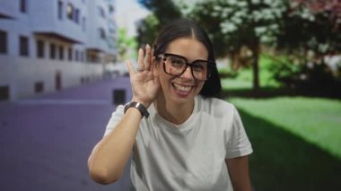 Kentin caddesinde bina ve ağaçlarla beyaz tişört giyen, el pençe divan duran, büyük gözlüklü, el pençe divan duran genç bir kadın..