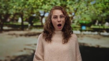 Gözlüklü bir kadın, arka planda yeşil yapraklı bir parkta el kol hareketi yaparken şaşkınlığını dile getiriyor..