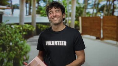 Sokakta elinde hediyeyle gülümseyen bir adam gönüllü tişört giyiyor. Etrafı yeşillik ve ağaçlarla çevrili. Güneşli bir günde pozitif bir atmosfer yaratıyor..
