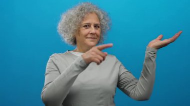 Mavi bir arka planda gri saçlı yaşlı bir kadın, bir eliyle işaret ediyor ve diğer eliyle avucunu açıyor, günlük gri giysiler giyiyor, izole ve anlamlı..