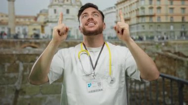 Doktor önlüğü ve steteskop giymiş sakallı genç adam, gülümsüyor ve Roma harabelerinin önünde yukarı doğru işaret ediyor..