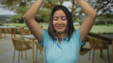 Mavi gömlekli kadın, güneşli bir restoran terasında kafasının üstünde kalp hareketi yapıyor. Arkasında tahta sandalyeler ve yeşil park var..